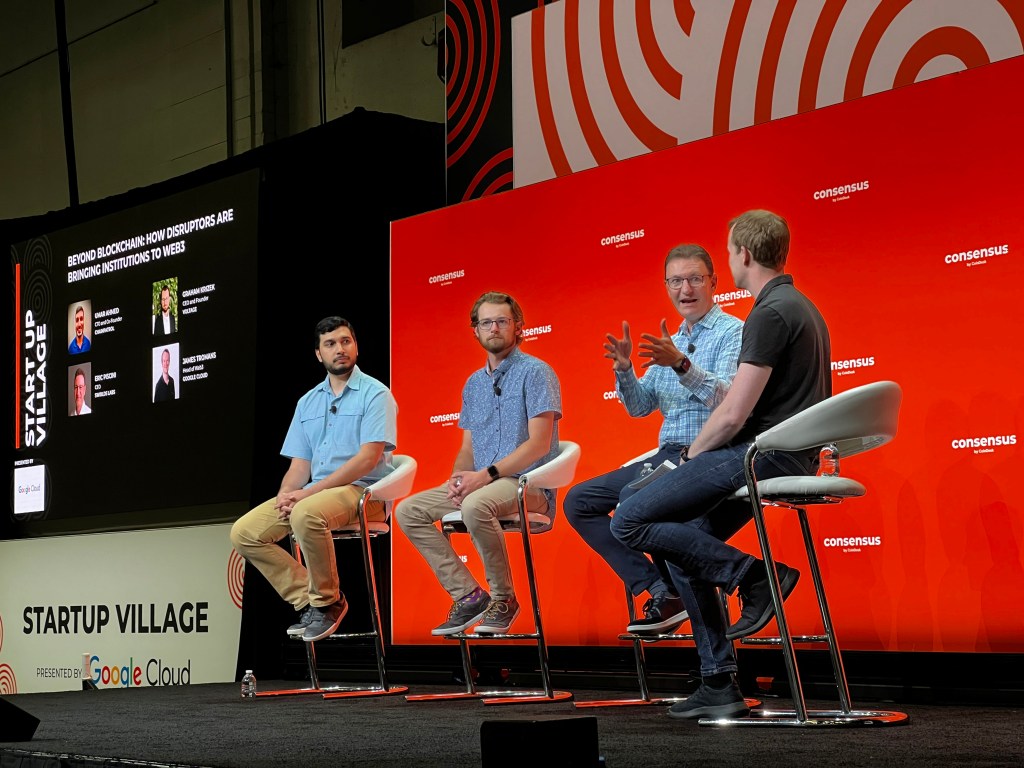 4 men sitting on chairs on a stage, for a panel at Consensus, with a red background behind them